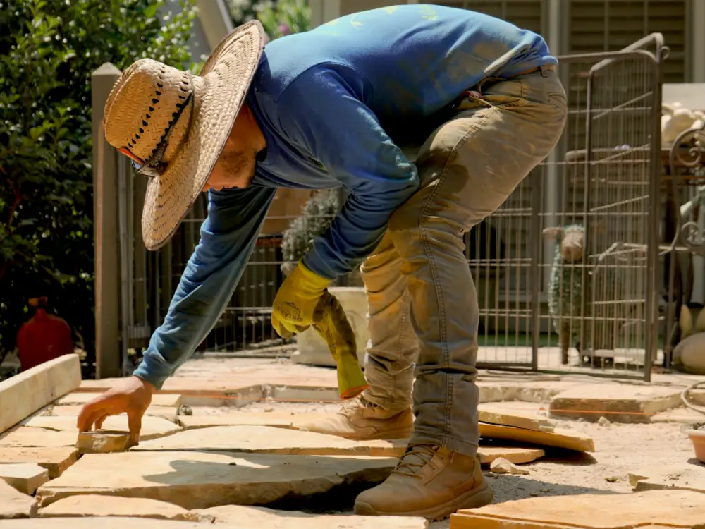 ALS crew member in a straw hat setting flagstone into a bedding layer by hand, cut pieces staged around him.