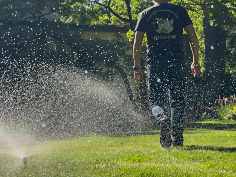 ALS crew member walking a lawn while a sprinkler head throws a wide arc of backlit spray across the grass.