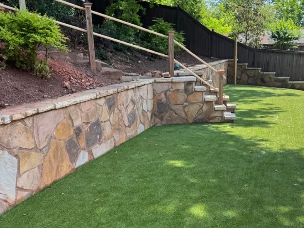 Finished stacked-stone retaining wall with steps and a rail, holding a graded slope above a level back lawn.